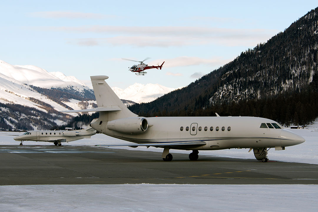 A red helicopter flying over two private jets at the airport of St Moritz, Switzerland.