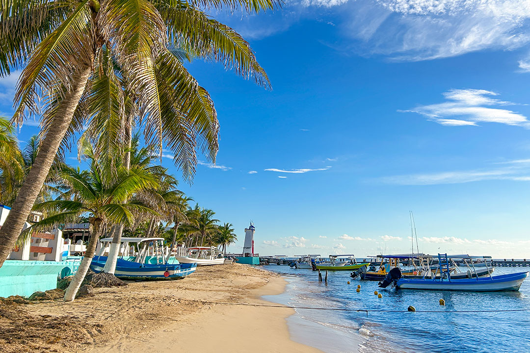 Riviera Maya beach with palm trees, boats and lighthouse on turquoise Caribbean sea.